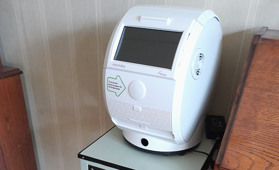 En bullig liten apparat står på ett avlastningsbord. På apparaten finns en pil med texten "Tryck på den gröna knappen för att få läkemedel". 